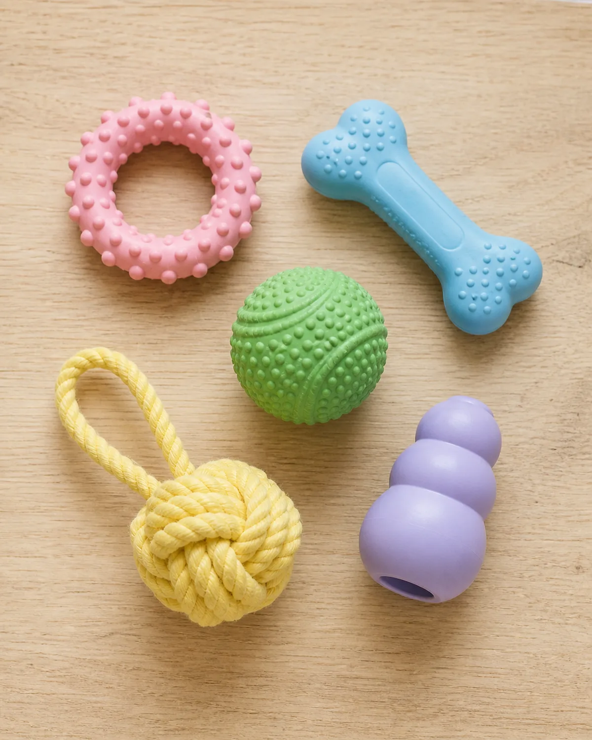 a flat lay on a light wood table showing five different puppy teething toys in soft rubber a pink nubbed ring a blue bone shape a green textured ball a yellow braided rope knot and a light purple puppy KONG all looking soft and squeezable in gentle natural window light from above