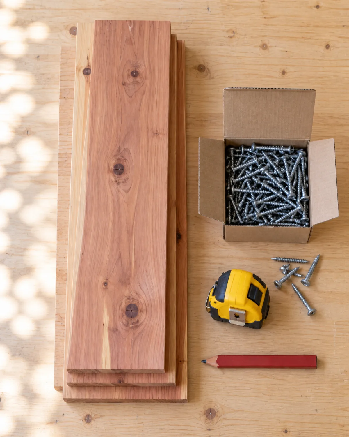 overhead view of stacked cedar planks and a box of galvanized deck screws and a tape measure and a carpenters pencil arranged on a pale workbench in a bright garage with natural light from an open garage door and clean organized workspace