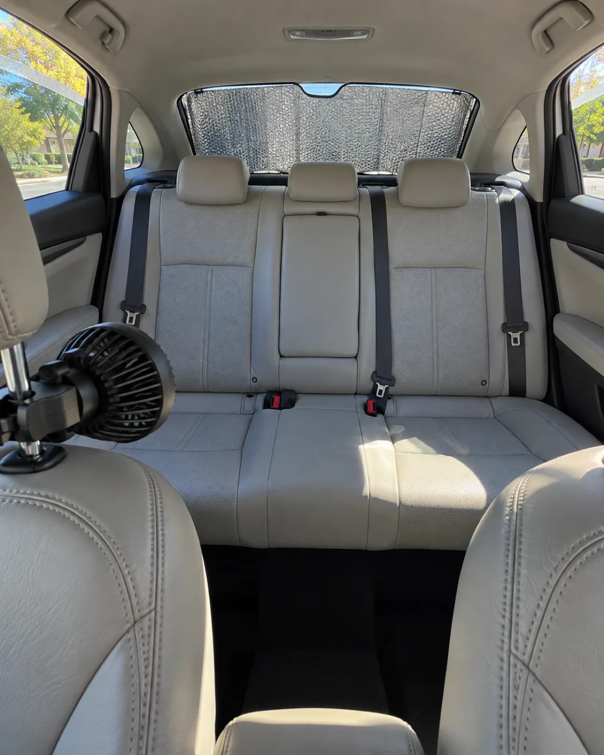 interior view of an empty car backseat with both rear windows cracked two inches and a clip-on fan attached to the headrest and a reflective sunshade on the rear window on a bright sunny day with pale leather seats