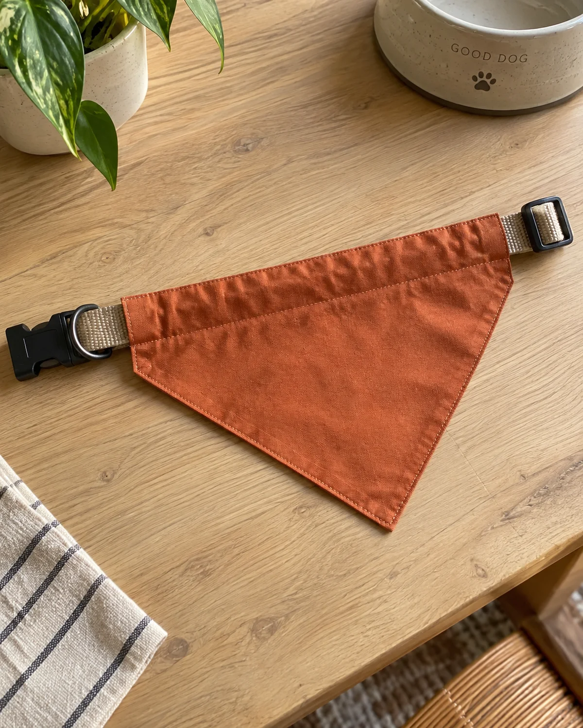 a finished triangle dog bandana in terracotta orange laid flat on a light wood table showing a neat fabric channel sewn along the top edge with a collar threaded through it